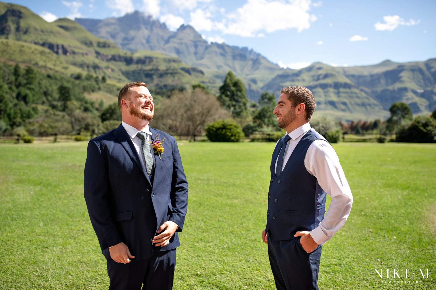 The groom sharing a joyful laugh with his best man on the lawn at Champagne Castle Hotel with the Central Drakensberg mountains rising behind them — a relaxed and genuine moment before the micro-wedding ceremony.