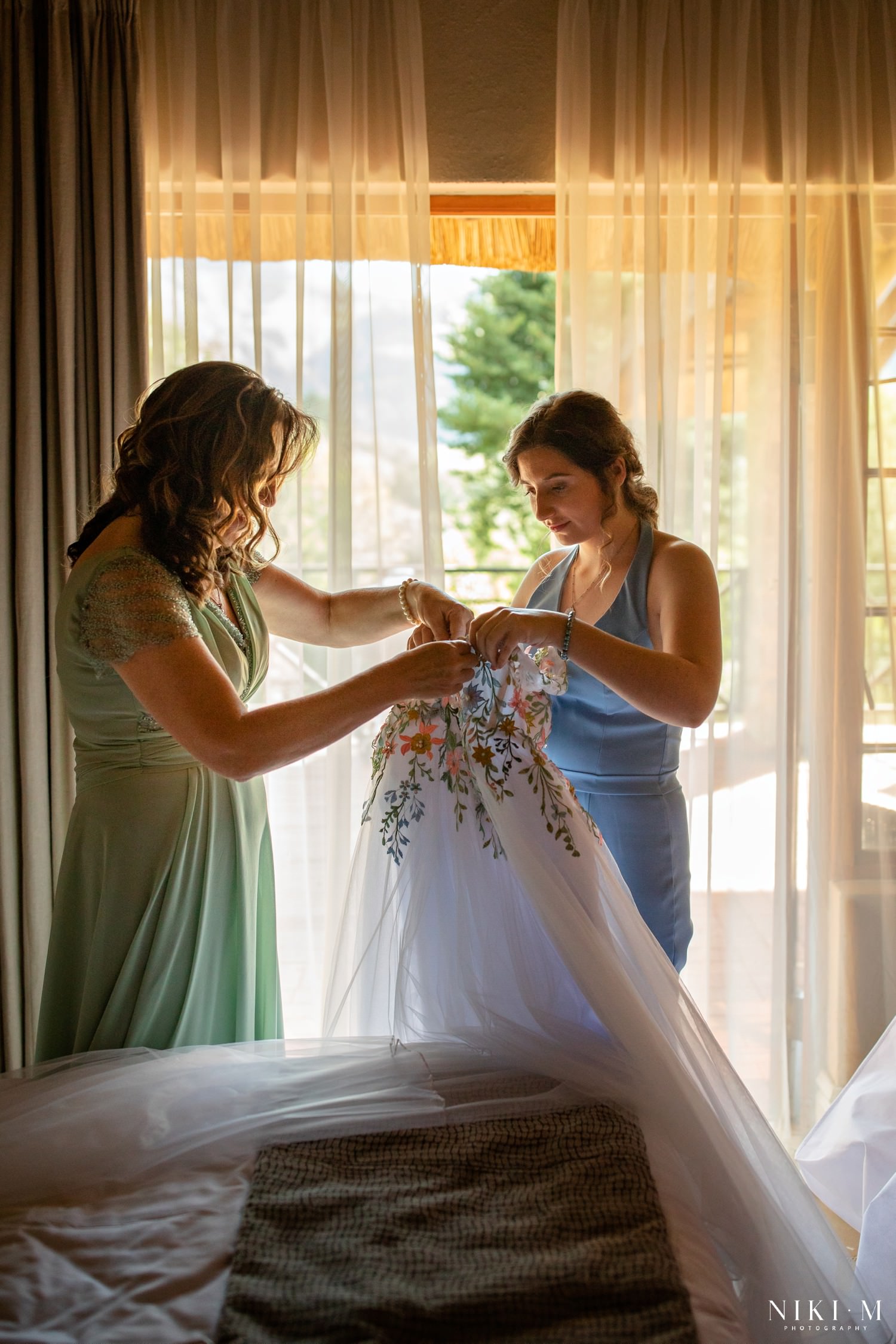 A mother and sister unbuttoning the bride's embroidered floral gown as golden sunlight filters through the curtains of Champagne Castle Hotel's bridal suite