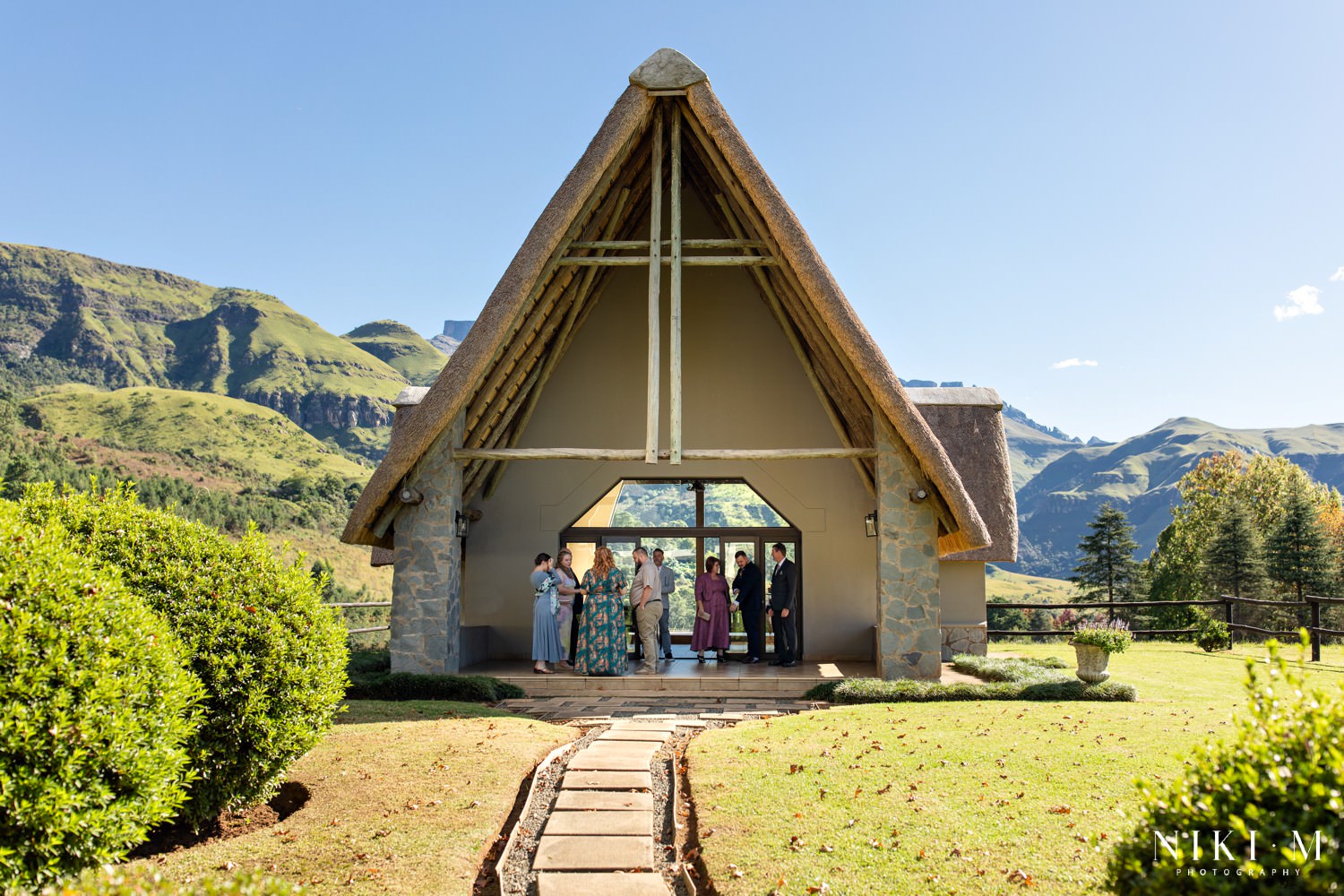 Guests gather outside the thatched chapel at Champagne Castle Hotel, surrounded by lush green peaks of the Central Drakensberg, ahead of a mountain wedding ceremony.