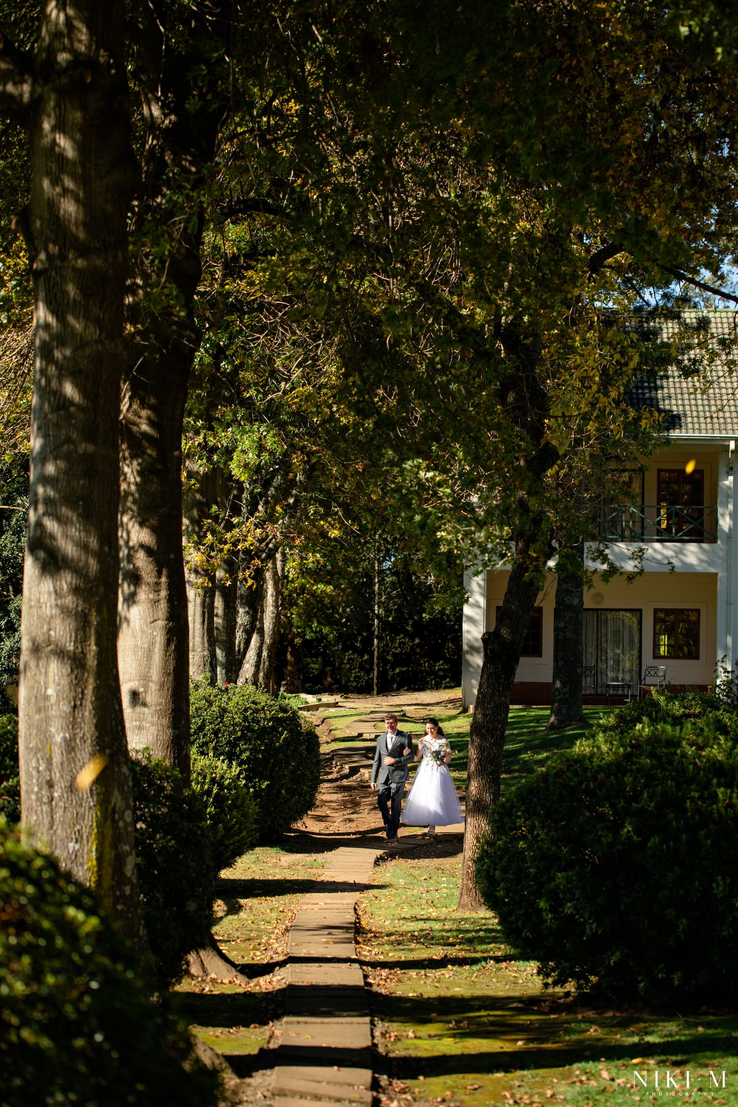 The bride walks arm-in-arm with her father down a tree-lined path toward the chapel, soft sunlight filtering through the canopy at Champagne Castle Hotel.
