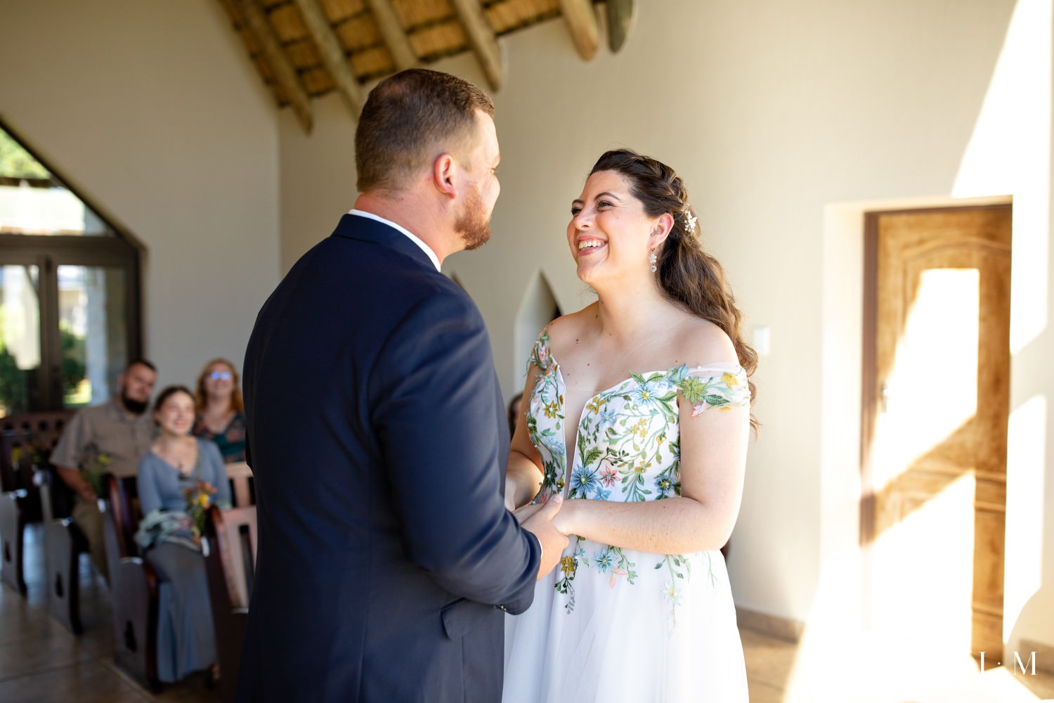 The bride and groom share a joyful smile during their intimate wedding ceremony at Champagne Castle Hotel’s glass-fronted chapel, sunlight streaming across the stone floor.