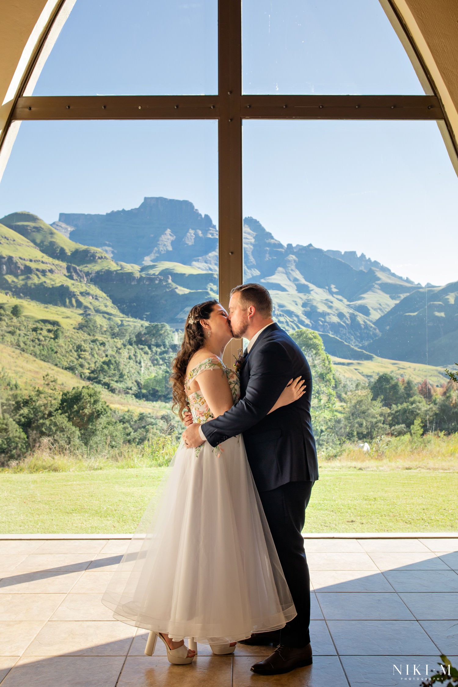 The couple share their first kiss framed by Champagne Castle’s breathtaking mountain view through the chapel window — a true Drakensberg wedding moment.