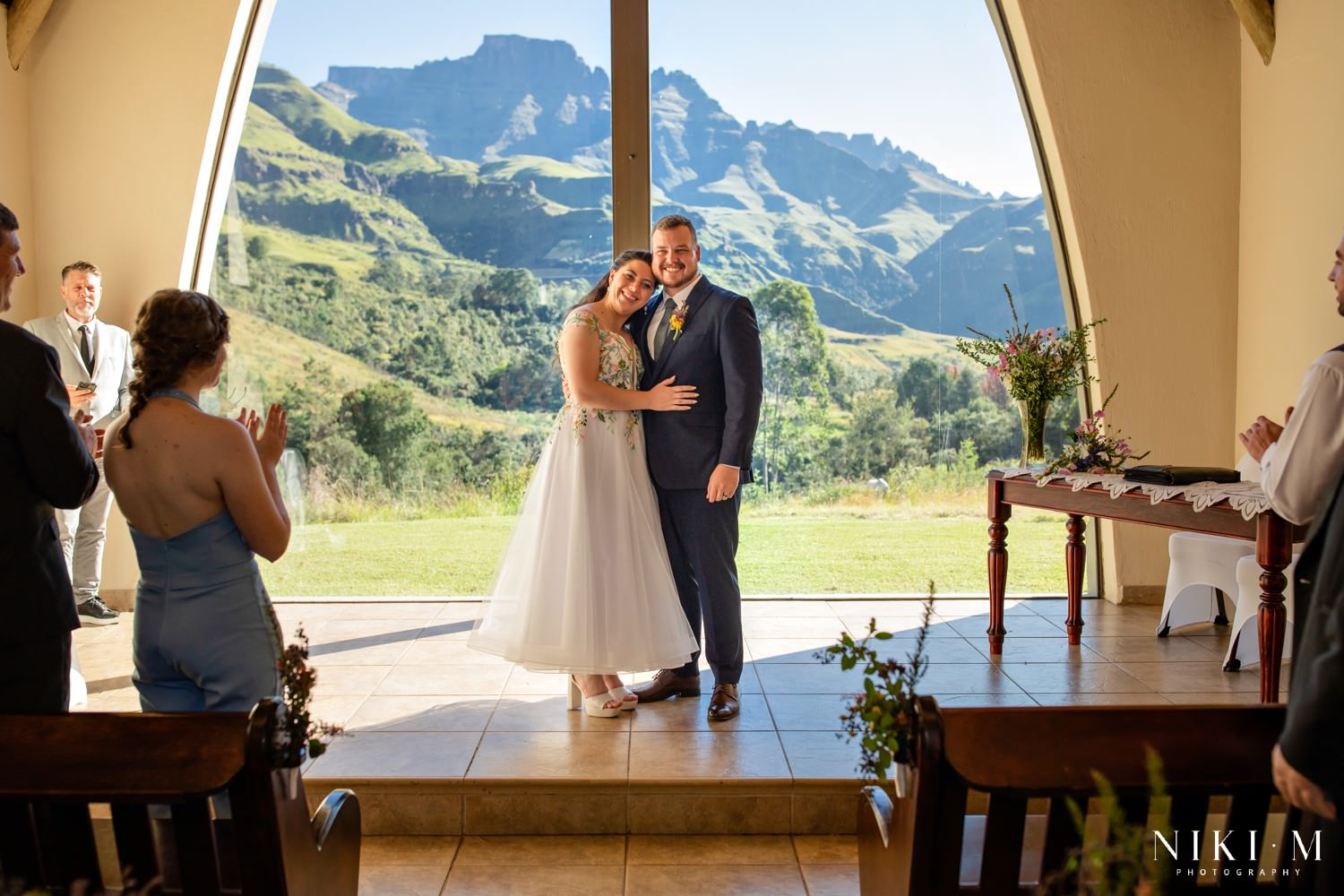 The newly married couple embrace and smile at guests, framed by Champagne Castle’s glass chapel and the rugged peaks of the Central Drakensberg.