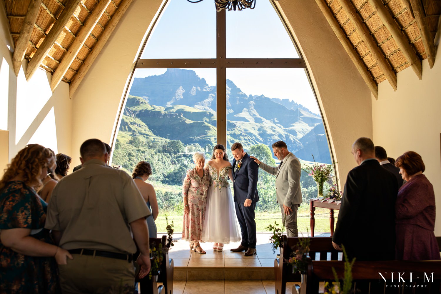 Wide shot of the entire ceremony group praying together at Champagne Castle Hotel, framed by the towering peaks of the Central Drakensberg beyond the chapel window.