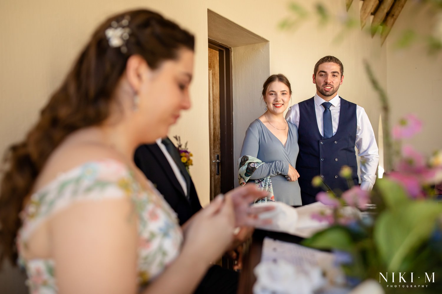 The bride signs the marriage register while family members look on proudly, surrounded by soft pastel florals and golden autumn light.