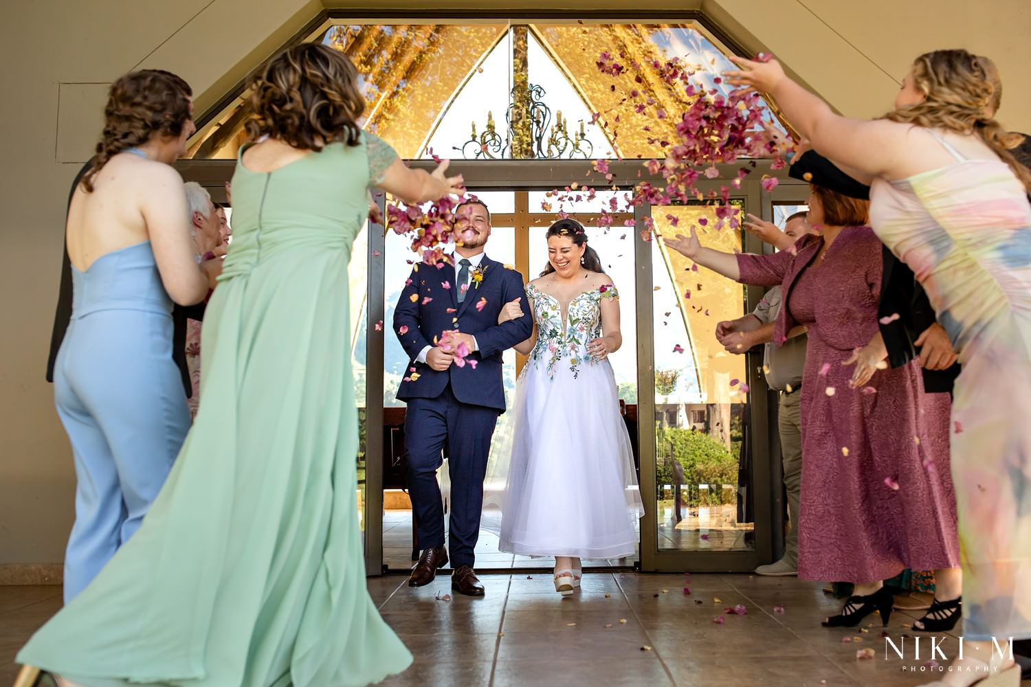 Guests cheer as the newlyweds walk down the aisle beneath a shower of rose petals, beaming as they exit the chapel into bright Drakensberg sunshine.