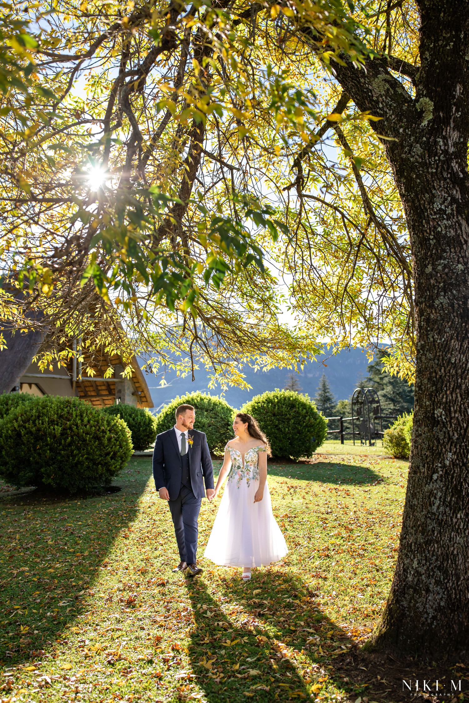 The wedding couple stroll hand in hand beneath golden autumn leaves at Champagne Castle Hotel, sunlight streaming through the trees during their romantic Drakensberg photo session.