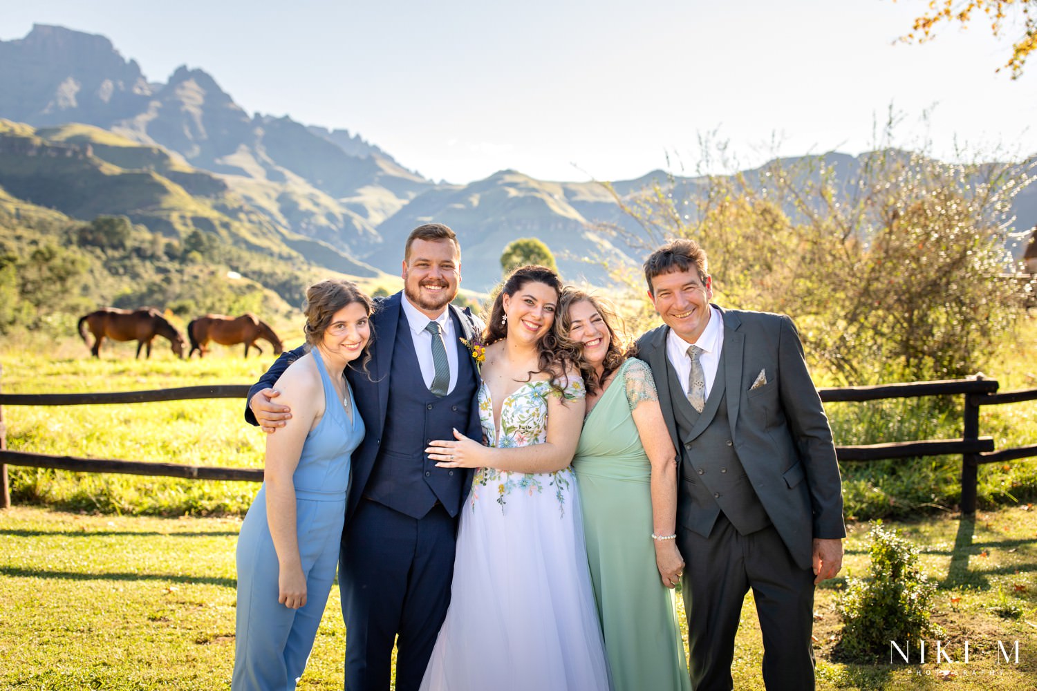 The bridal couple and family smile proudly against the mountain backdrop of Champagne Castle Hotel, a natural and joyful Central Drakensberg family portrait.
