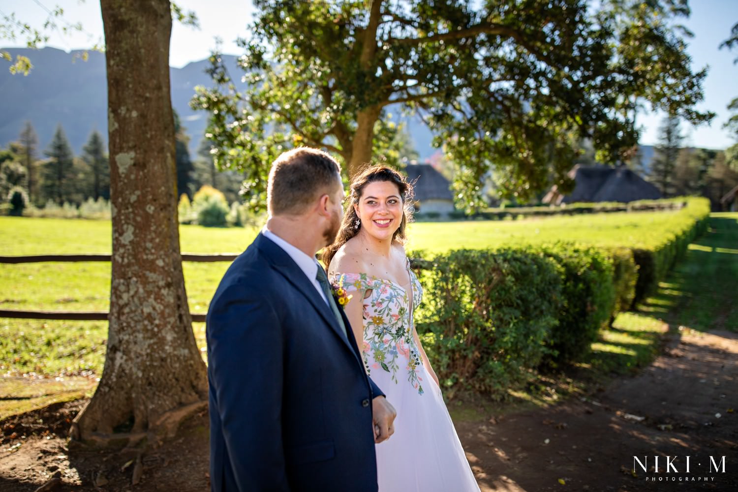 Bride and groom walking together under golden afternoon light at Champagne Castle Hotel gardens with Drakensberg peaks in the background.