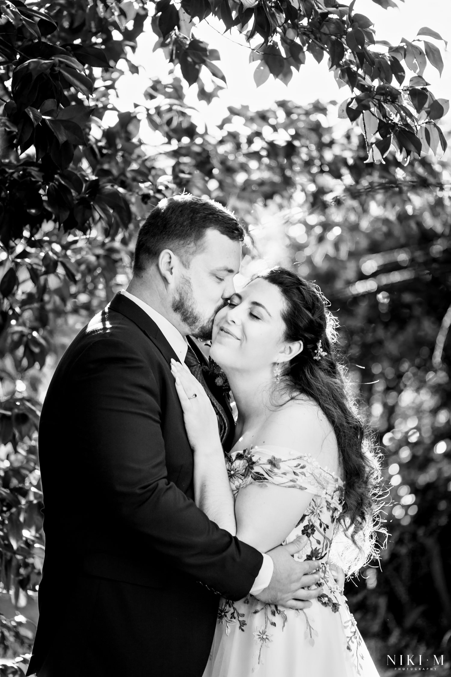 Black and white portrait of the couple sharing a quiet embrace beneath leafy branches. A tender Champagne Castle Hotel wedding moment.
