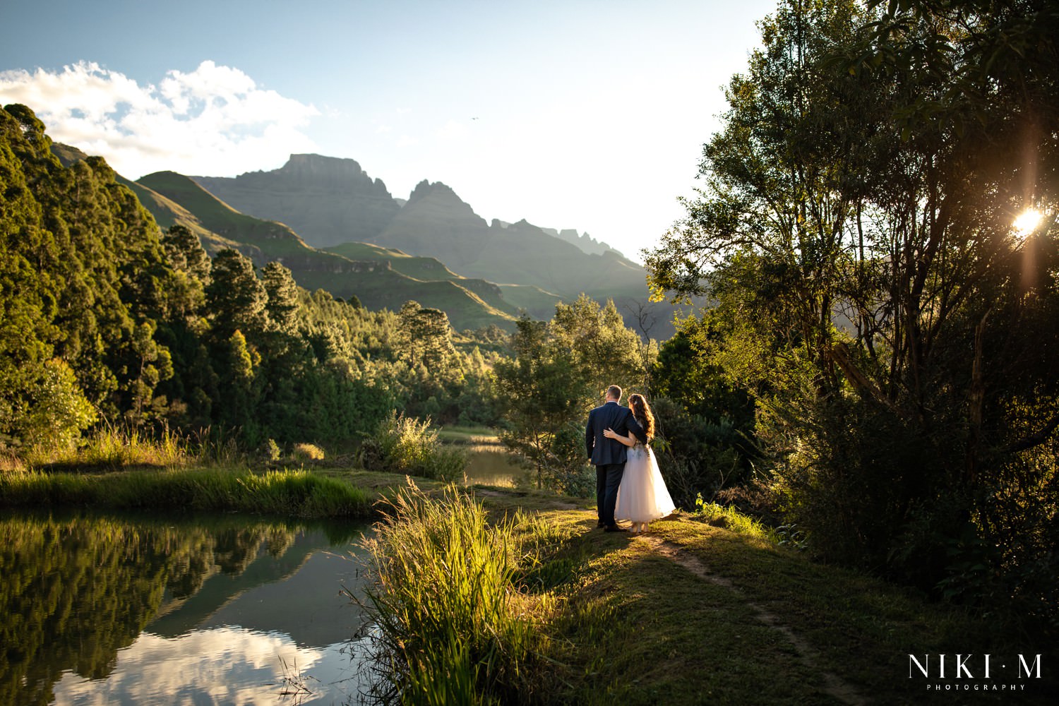 Scenic Drakensberg mountain wedding photo showing the couple walking away arm in arm beside a still pond reflecting the Cathedral Peak range in the late afternoon light.