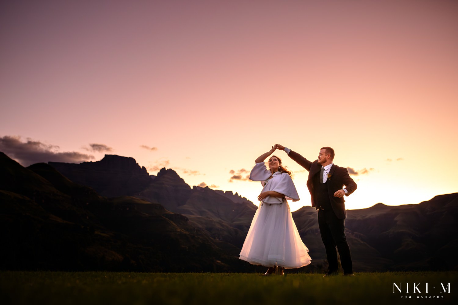 Couple dancing at sunset with Champagne Castle silhouetted in the background, the sky painted pink and gold — a breathtaking moment in the heart of the Central Drakensberg.