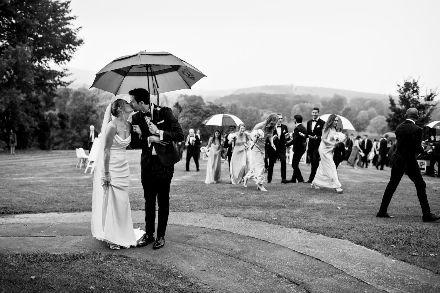 A joyful and frantic end to a wedding ceremony at one of the top Drakensberg wedding venues, Champagne Sports Resort, with the bride and groom sharing a kiss under an umbrella as their guests laugh and walk behind them in the pouring rain. The mountains and classic black-and-white tones capture the romance and timeless charm of a Drakensberg celebration.