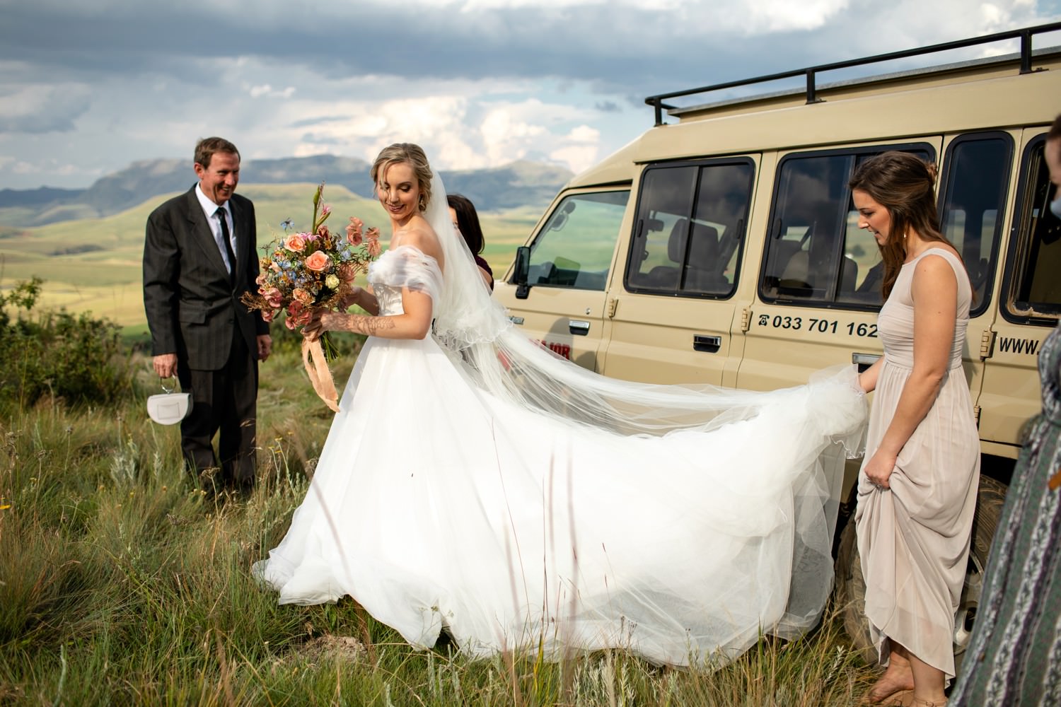 Bride stepping gracefully from a safari-style vehicle at her mountaintop micro wedding in the Southern Drakensberg, helped by her bridesmaids and father. The sweeping mountain views, soft light, and elegant gown highlight the adventurous spirit and natural beauty that make the Drakensberg so unforgettable as a wedding venue.
