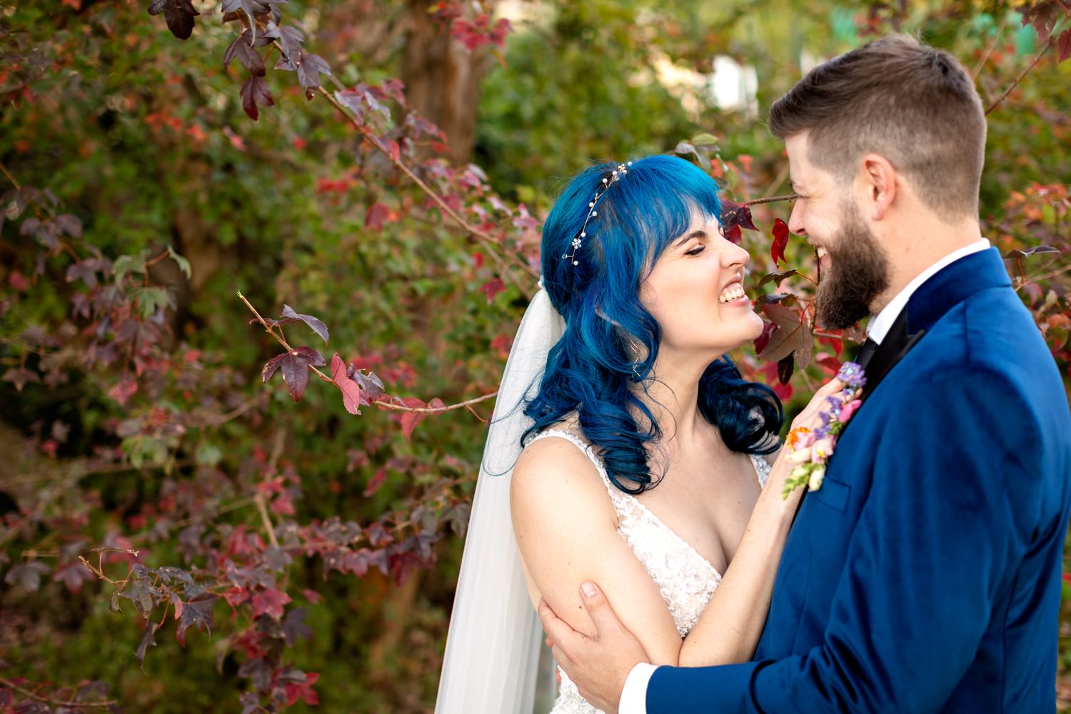 Bride with vibrant blue hair and groom in a navy velvet suit share an intimate moment among autumn foliage at at Drakensberg wedding venue Dragon Peaks Resort. The colourful tones and natural setting show how unique and personal mountain weddings can be.