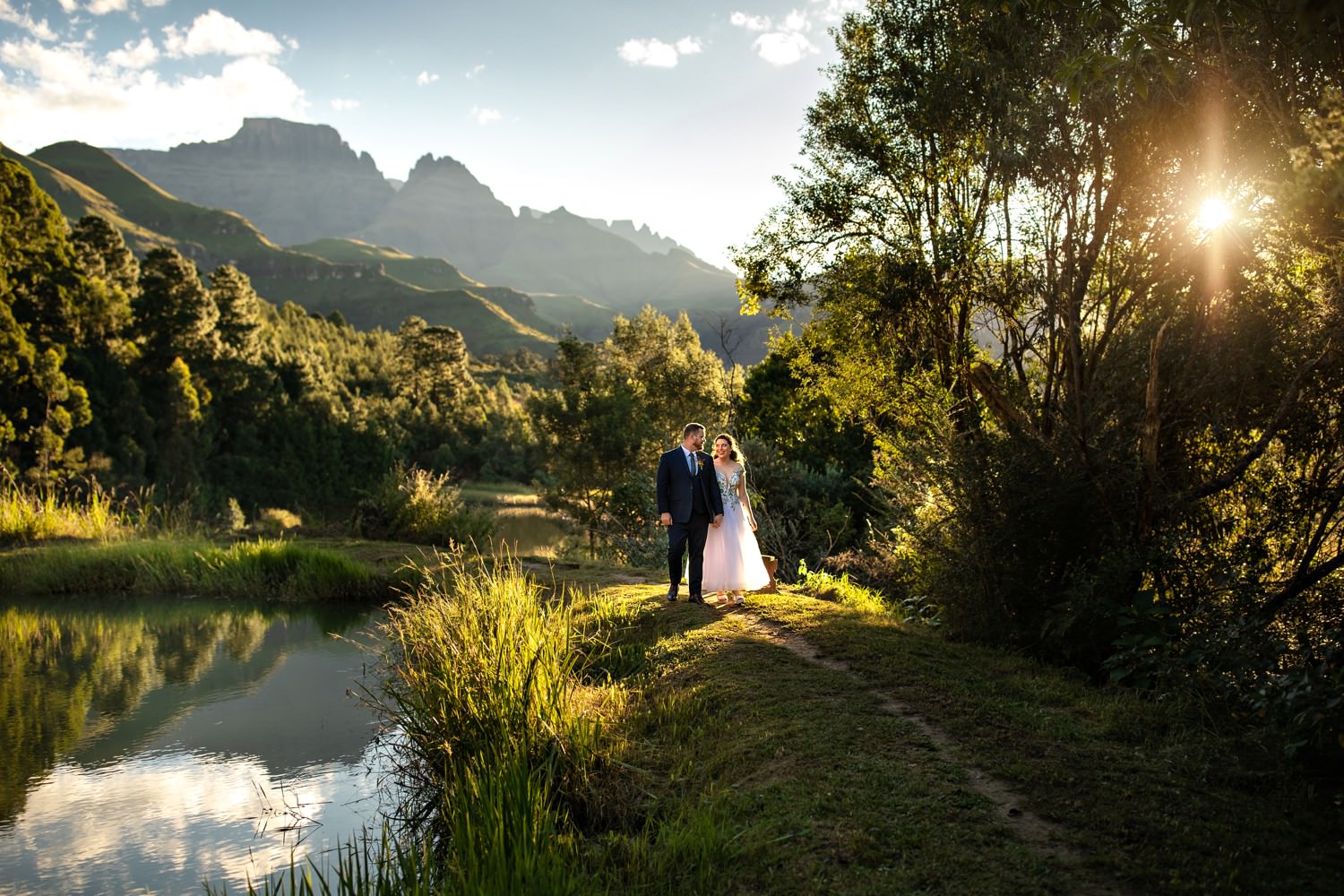 Bride and groom stroll hand-in-hand beside a still mountain pond framed by lush greenery at Champagne Castle Hotel, reflecting the tranquil atmosphere of Drakensberg wedding venues. The golden afternoon light and dramatic peaks in the distance capture the serenity and romance of a mountain wedding.