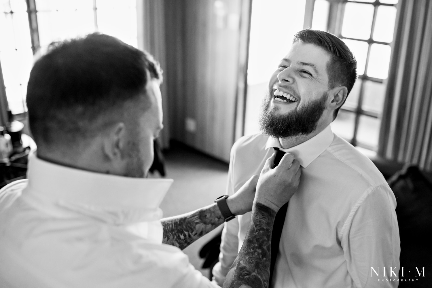 Groomsman adjusting groom's tie while he laughs, black and white portrait at a Drakensberg wedding