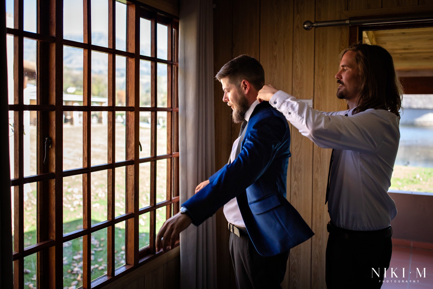 Groom putting on his blue velvet jacket at a window with the Drakensberg mountains visible behind him