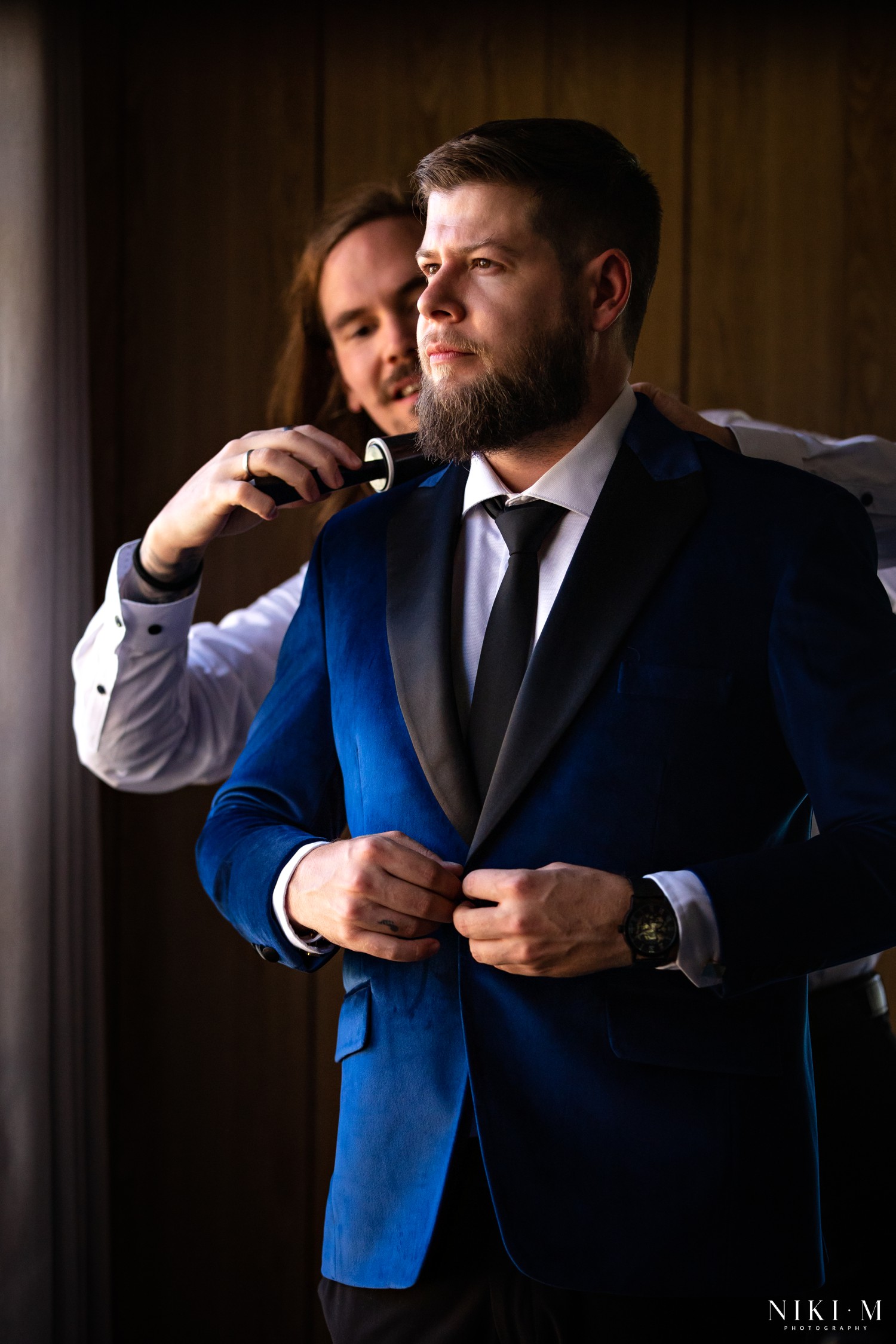 Groom in blue velvet tuxedo jacket being lint-rolled in dramatic window light at a Drakensberg wedding