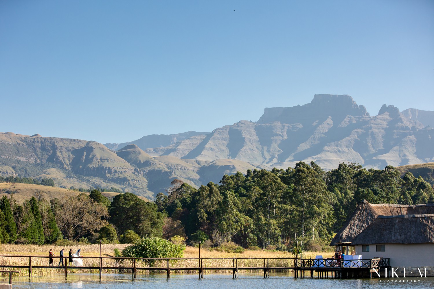 Bridal party crossing a wooden jetty with the Drakensberg escarpment behind them, photographed by Drakensberg wedding photographer Niki M