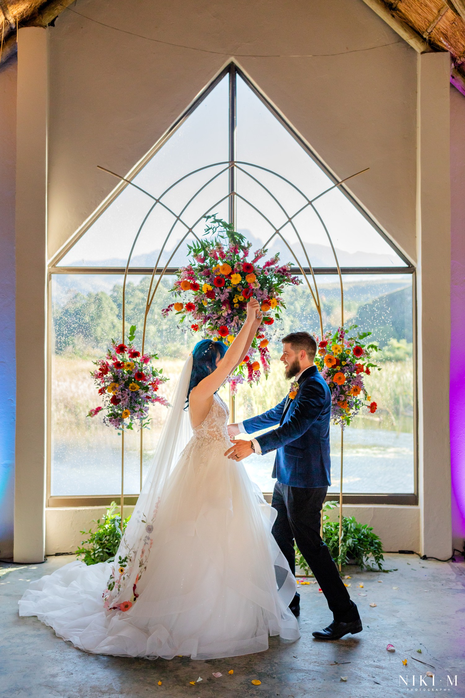 Newlyweds celebrating beneath a gold wildflower arch with the Drakensberg mountains through the window behind them