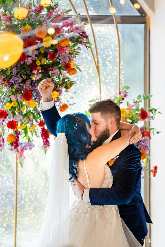 First kiss with the groom's fist raised in celebration beneath the floral arch at a Champagne Valley wedding