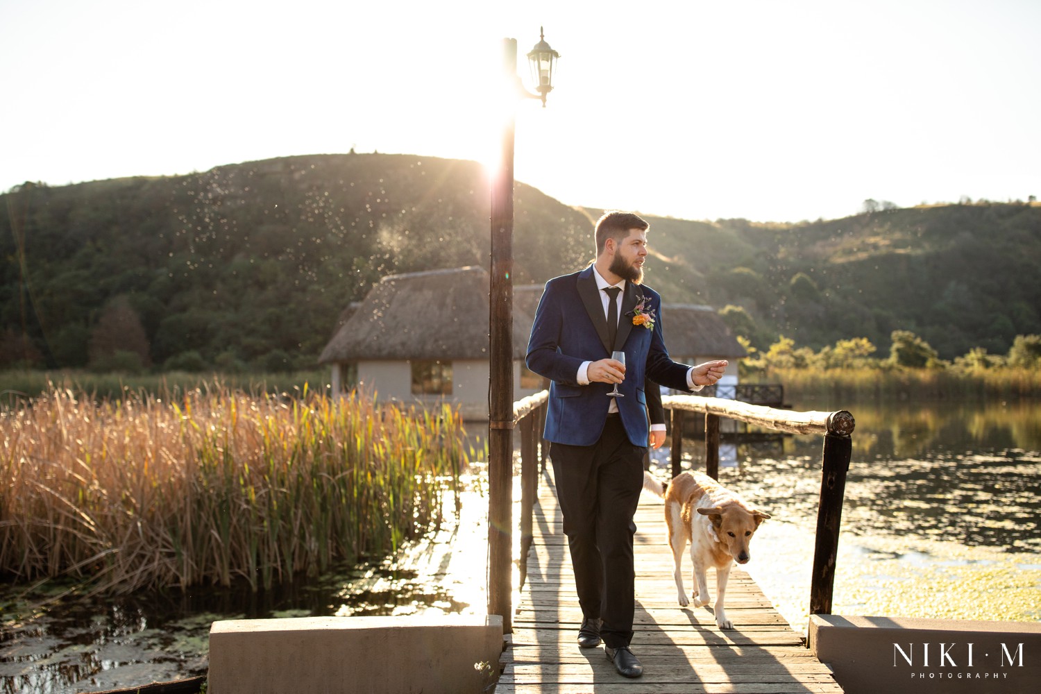 Groom in blue velvet jacket walking the jetty with dog Sadie at golden hour, photographed by Drakensberg wedding photographer Niki M