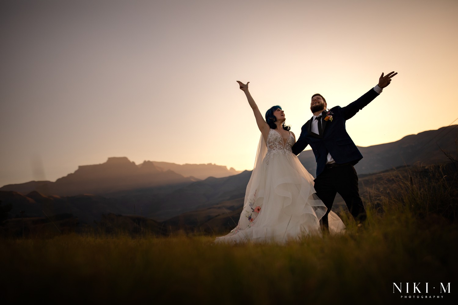 Bride and groom with arms raised as the Drakensberg escarpment glows at dusk behind them