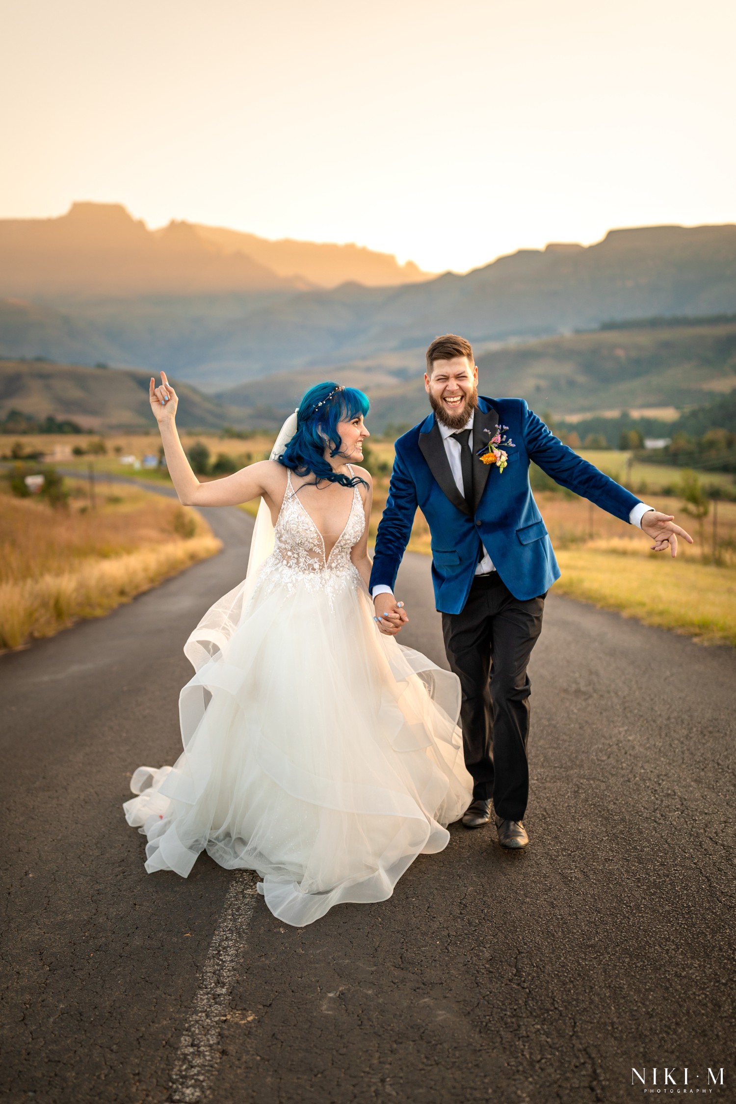 Bride and groom walking the open road at golden hour with the Drakensberg escarpment glowing behind them, photographed by Drakensberg wedding photographer Niki M