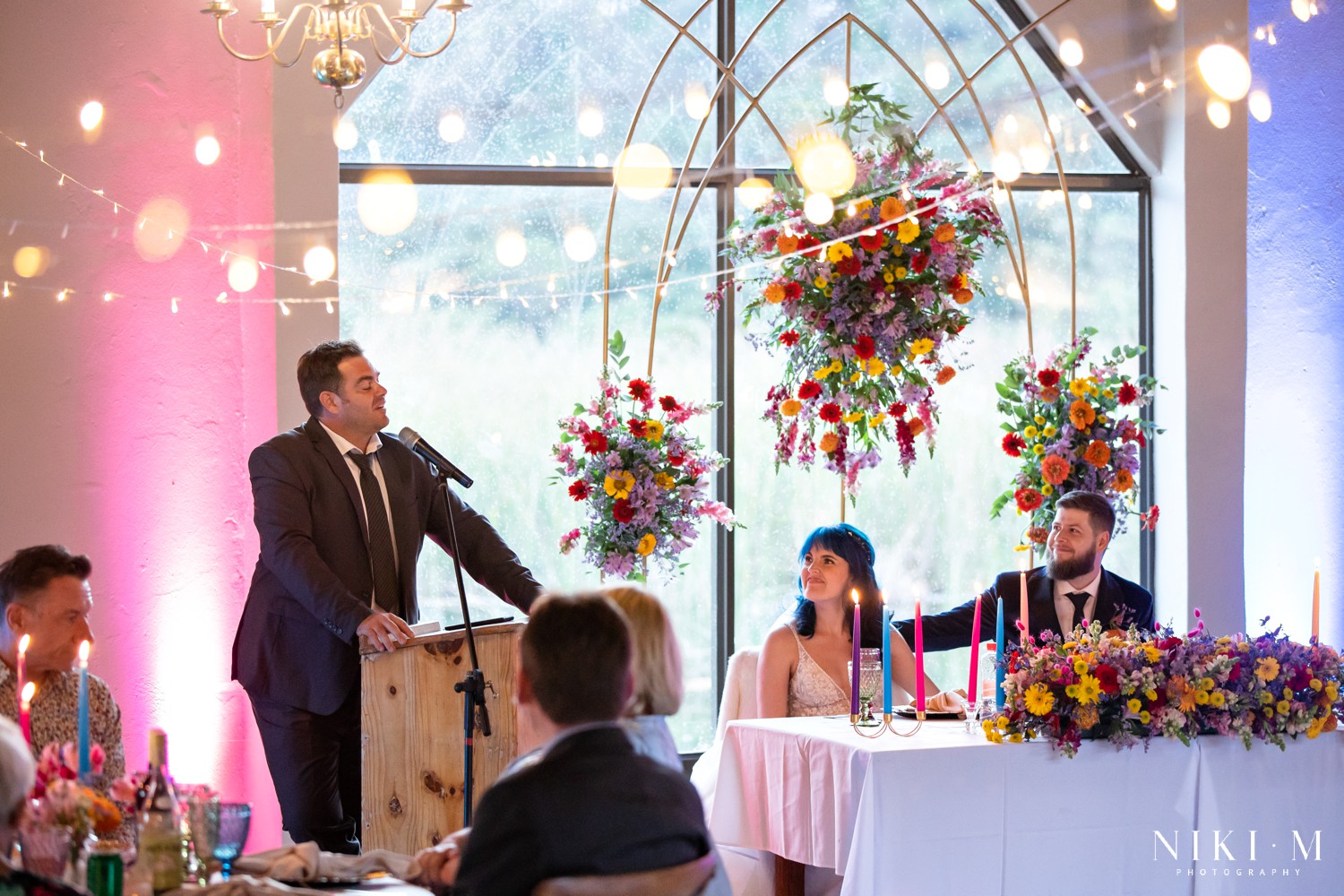 Guest giving a speech with the couple beneath a hanging wildflower installation at a Champagne Valley Drakensberg wedding